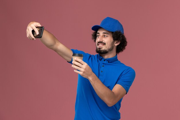 Vista frontal del mensajero masculino en uniforme azul y gorra con taza de café de entrega en sus manos y tomando una fotografía en la pared rosa