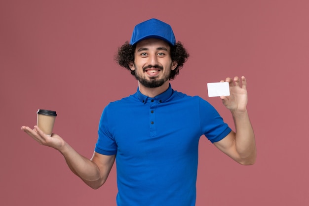 Foto gratuita vista frontal del mensajero masculino en uniforme azul y gorra con taza de café de entrega y tarjeta en sus manos en la pared rosa