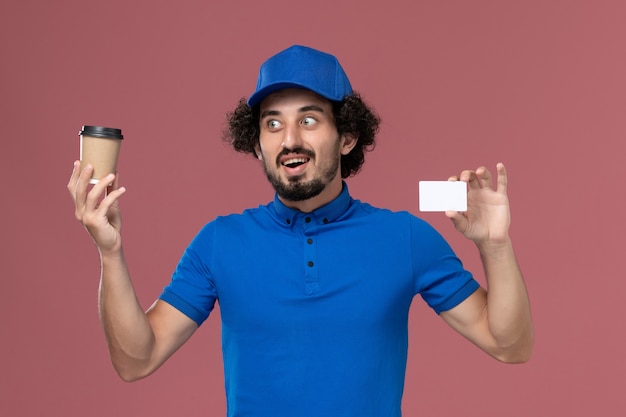 Vista frontal del mensajero masculino en uniforme azul y gorra con taza de café de entrega y tarjeta en sus manos en la pared rosa