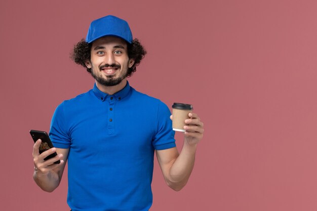Vista frontal del mensajero masculino en uniforme azul y gorra con taza de café de entrega y teléfono inteligente en sus manos en la pared rosa