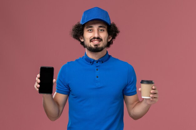 Vista frontal del mensajero masculino en uniforme azul y gorra con taza de café de entrega y teléfono en sus manos en la pared rosa