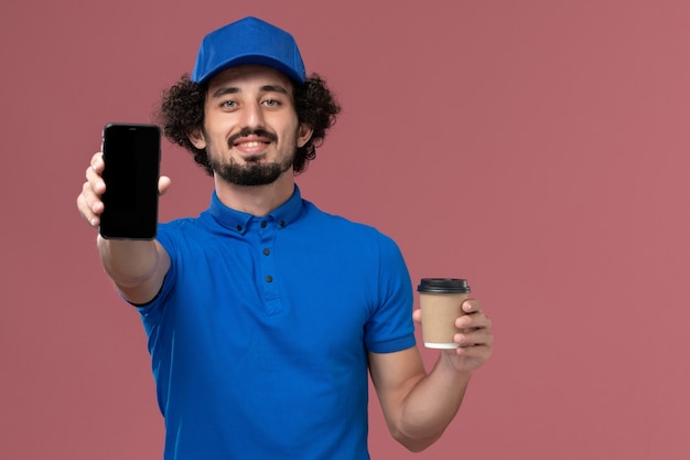Vista frontal del mensajero masculino en uniforme azul y gorra con taza de café de entrega y teléfono en sus manos en la pared rosa
