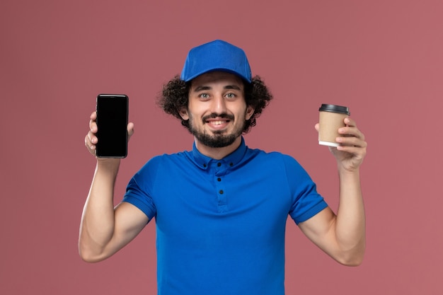 Vista frontal del mensajero masculino en uniforme azul y gorra con taza de café de entrega y teléfono en sus manos en la pared rosa