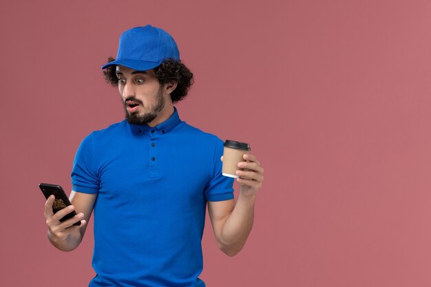 Vista frontal del mensajero masculino en uniforme azul y gorra con taza de café de entrega y teléfono en sus manos en la pared rosa