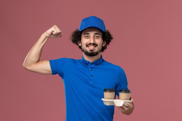 Vista frontal del mensajero masculino en uniforme azul y gorra con tazas de café de entrega en sus manos flexionando en la pared rosa