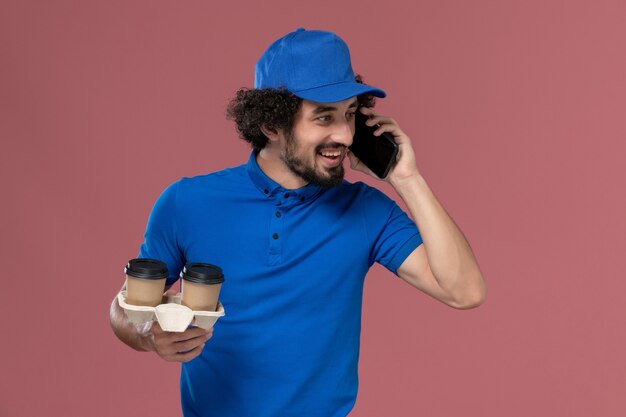 Vista frontal del mensajero masculino en uniforme azul y gorra con tazas de café de entrega en sus manos hablando por teléfono en la pared rosa