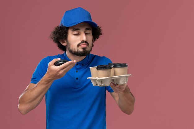 Vista frontal del mensajero masculino en uniforme azul y gorra con tazas de café de entrega en sus manos con olor a fragancia en la pared rosa claro