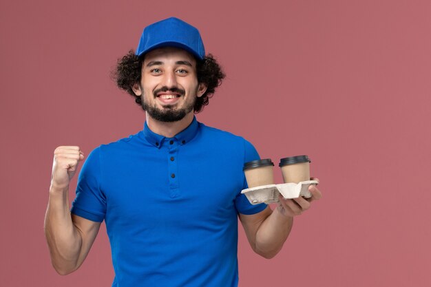 Vista frontal del mensajero masculino en uniforme azul y gorra con tazas de café de entrega en sus manos en la pared rosa claro