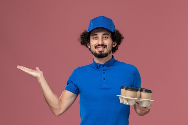 Vista frontal del mensajero masculino en uniforme azul y gorra con tazas de café de entrega en sus manos en la pared rosa claro