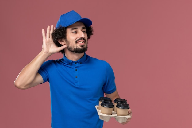 Foto gratuita vista frontal del mensajero masculino en uniforme azul y gorra con tazas de café de entrega en sus manos en la pared rosa claro