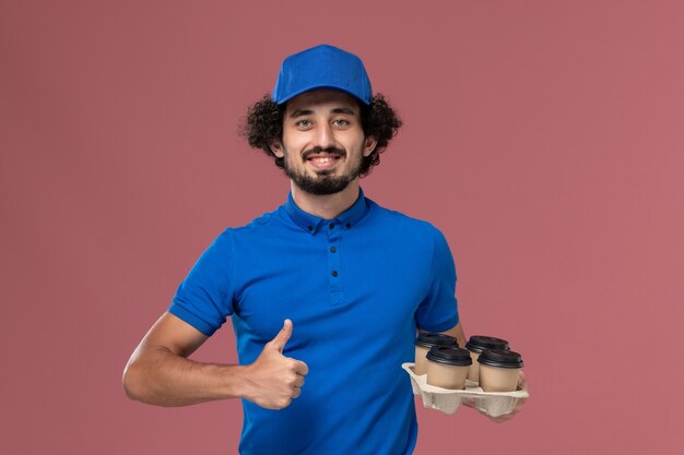 Vista frontal del mensajero masculino en uniforme azul y gorra con tazas de café de entrega en sus manos en la pared rosa claro