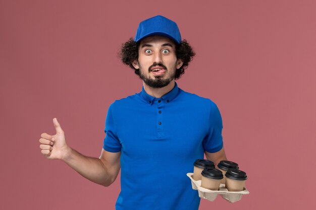 Vista frontal del mensajero masculino en uniforme azul y gorra con tazas de café de entrega en sus manos en la pared rosa claro