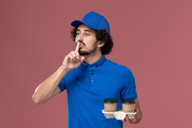 Vista frontal del mensajero masculino en uniforme azul y gorra con tazas de café de entrega en sus manos en la pared rosa