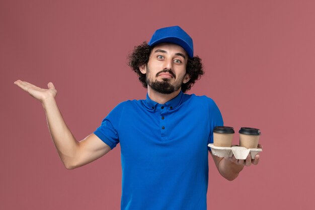 Vista frontal del mensajero masculino en uniforme azul y gorra con tazas de café de entrega en sus manos en la pared rosa