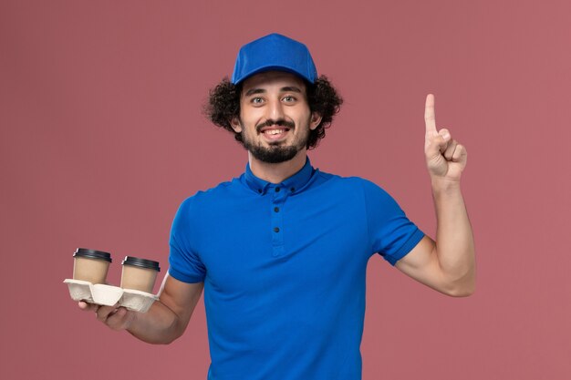 Vista frontal del mensajero masculino en uniforme azul y gorra con tazas de café de entrega en sus manos en la pared rosa
