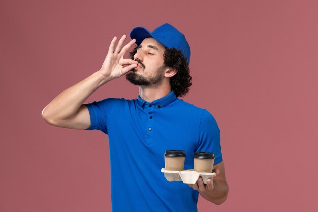 Vista frontal del mensajero masculino en uniforme azul y gorra con tazas de café de entrega en sus manos en la pared rosa