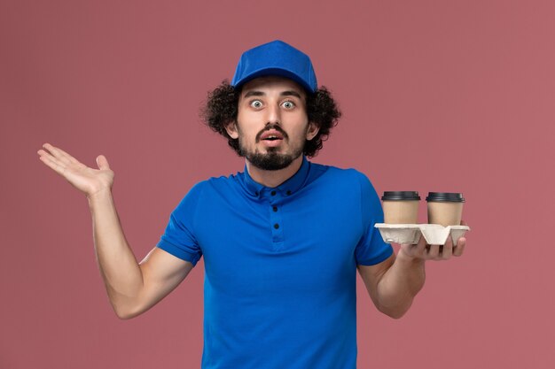 Vista frontal del mensajero masculino en uniforme azul y gorra con tazas de café de entrega en sus manos en la pared rosa