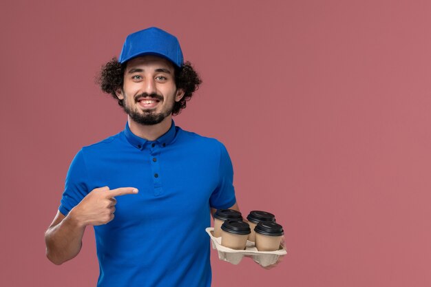 Vista frontal del mensajero masculino en uniforme azul y gorra con tazas de café de entrega en sus manos en la pared rosa