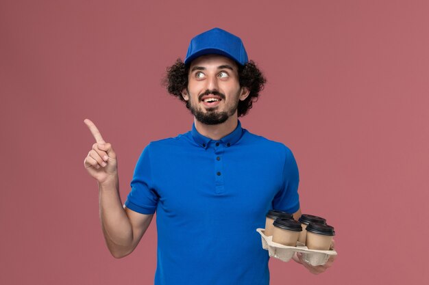 Vista frontal del mensajero masculino en uniforme azul y gorra con tazas de café de entrega en sus manos pensando en la pared rosa