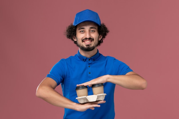 Vista frontal del mensajero masculino en uniforme azul y gorra con tazas de café de entrega en sus manos sonriendo en la pared rosa