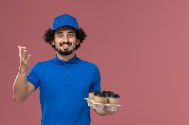 Vista frontal del mensajero masculino en uniforme azul y gorra con tazas de café de entrega en sus manos sonriendo en la pared rosa