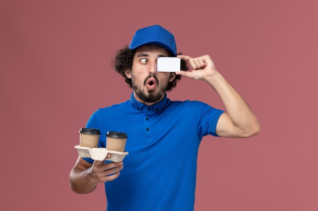 Vista frontal del mensajero masculino en uniforme azul y gorra con tazas de café de entrega y tarjeta en sus manos en la pared rosa
