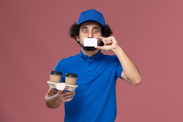 Vista frontal del mensajero masculino en uniforme azul y gorra con tazas de café de entrega y tarjeta en sus manos en la pared rosa