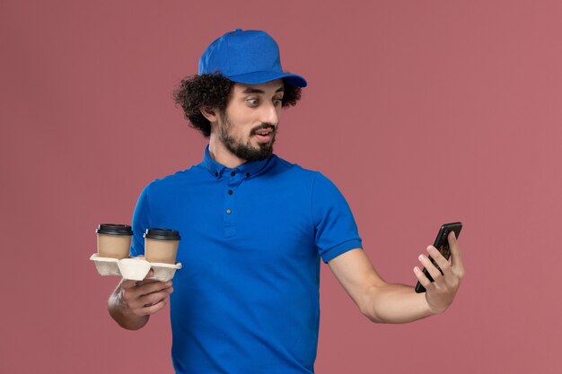 Vista frontal del mensajero masculino en uniforme azul y gorra con tazas de café de entrega y teléfono de trabajo en sus manos en la pared rosa