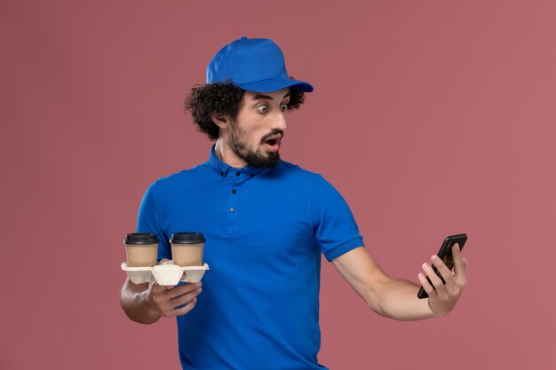 Vista frontal del mensajero masculino en uniforme azul y gorra con tazas de café de entrega y teléfono de trabajo en sus manos en la pared rosa