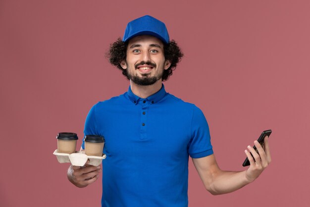 Vista frontal del mensajero masculino en uniforme azul y gorra con tazas de café de entrega y teléfono de trabajo en sus manos en la pared rosa