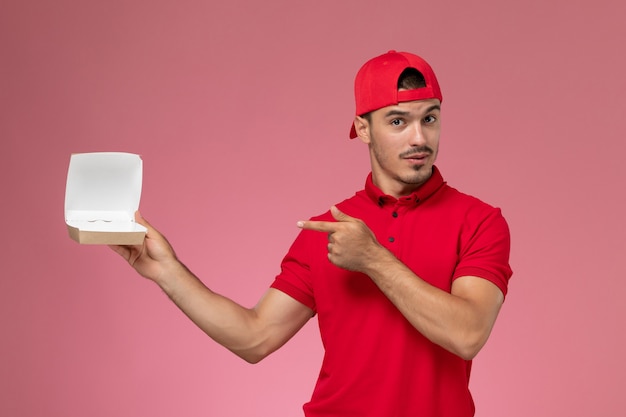 Vista frontal del mensajero masculino en uniforme rojo y gorra con paquete de entrega pequeño vacío en la pared rosa