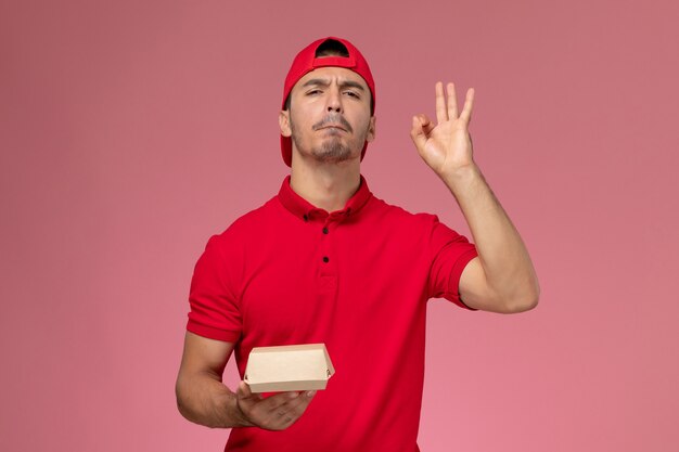 Vista frontal del mensajero masculino en uniforme rojo y gorra sosteniendo un pequeño paquete de entrega en la pared rosa