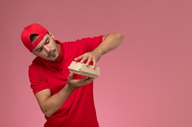 Foto gratuita vista frontal del mensajero masculino en uniforme rojo y gorra sosteniendo un pequeño paquete de entrega en la pared rosa