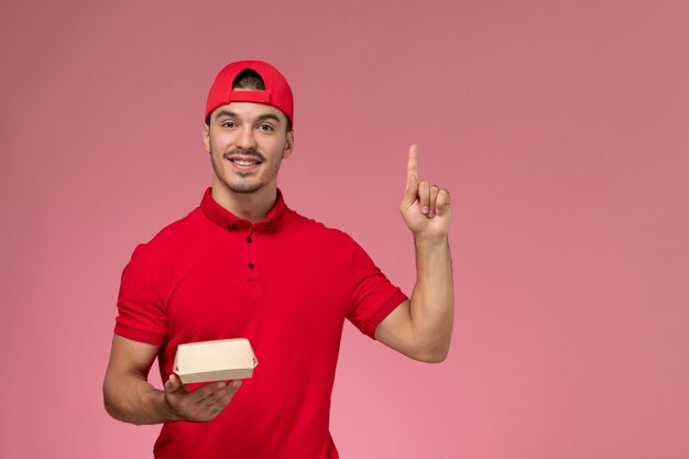 Vista frontal del mensajero masculino en uniforme rojo y gorra sosteniendo un pequeño paquete de entrega en la pared rosa