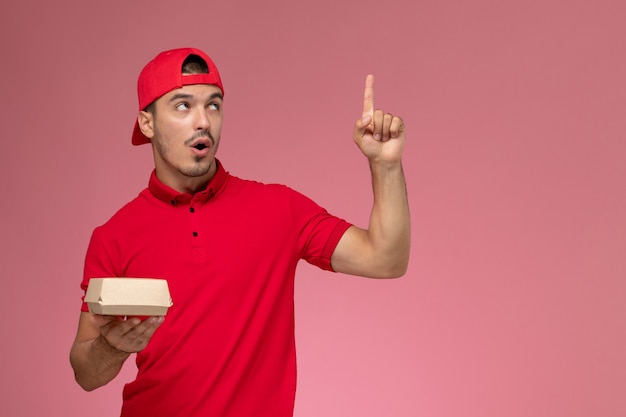 Vista frontal del mensajero masculino en uniforme rojo y gorra sosteniendo un pequeño paquete de entrega en la pared rosa