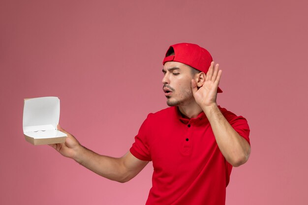 Vista frontal del mensajero masculino en uniforme rojo y gorra sosteniendo un pequeño paquete de entrega y tratando de escuchar en la pared rosa
