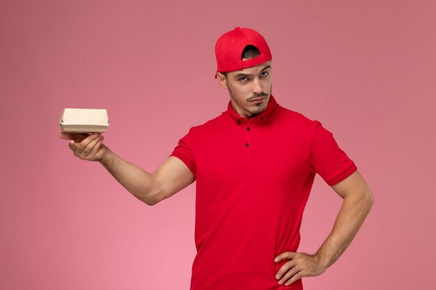 Vista frontal del mensajero masculino en uniforme rojo y gorra sosteniendo un pequeño paquete de entrega