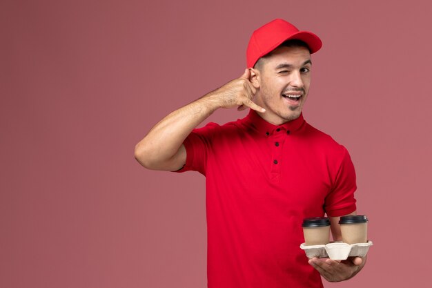 Vista frontal mensajero masculino en uniforme rojo sosteniendo tazas de café de entrega marrón guiñando un ojo en rosa macho trabajador de pared