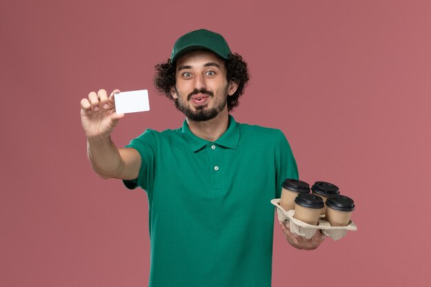 Vista frontal mensajero masculino en uniforme verde y capa sosteniendo tazas de café con tarjeta en servicio de escritorio rosa uniforme de trabajo trabajador de entrega masculino