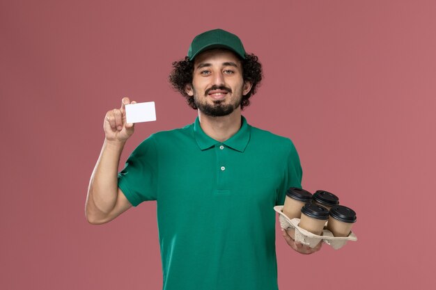 Vista frontal mensajero masculino en uniforme verde y capa sosteniendo tazas de café con tarjeta sobre fondo rosa servicio de entrega uniforme trabajador masculino