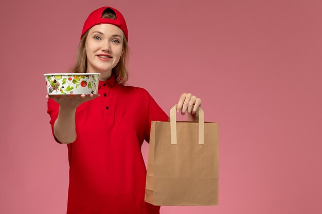 Vista frontal, mensajero, mujer joven, en, uniforme rojo, y, capa, tenencia, entrega, paquete de comida, y, tazón, sonriente, en, pared rosa