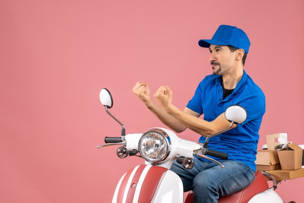 Vista frontal del mensajero nervioso chico con sombrero sentado en scooter sobre fondo de melocotón pastel