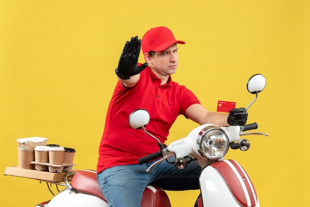 Vista frontal del mensajero nervioso hombre vestido con blusa roja y guantes de sombrero en máscara médica entregando orden sentado en scooter mostrando tarjeta bancaria