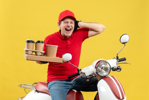 Vista frontal del mensajero nervioso hombre vestido con blusa roja y guantes de sombrero en máscara médica entregando orden sentado en scooter sosteniendo órdenes cerrando su oreja