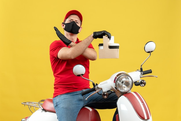 Vista frontal del mensajero nervioso hombre vestido con blusa roja y guantes de sombrero en máscara médica entregando orden sentado en scooter sosteniendo órdenes haciendo gesto de parada