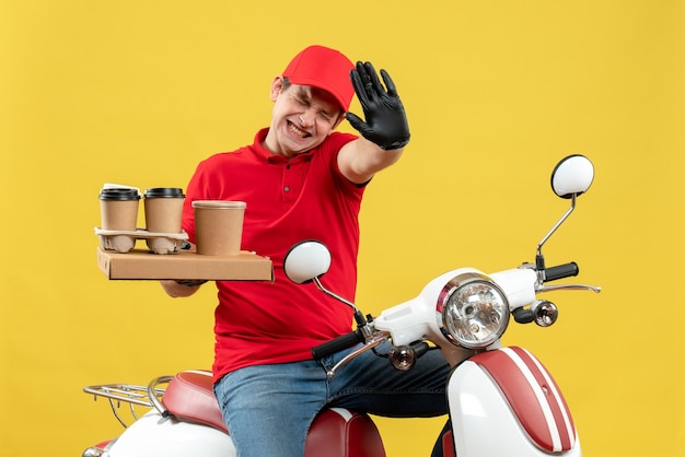 Vista frontal del mensajero nervioso preocupado vestido con blusa roja y guantes de sombrero en máscara médica entregando orden sentado en scooter con órdenes