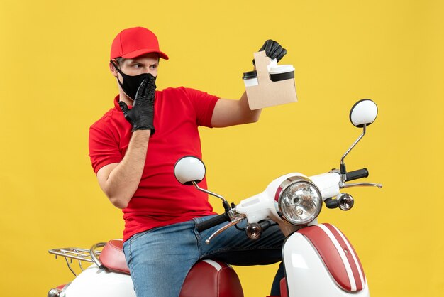 Vista frontal del mensajero ocupado enfocado sorprendido hombre vestido con blusa roja y guantes de sombrero en máscara médica sentado en scooter