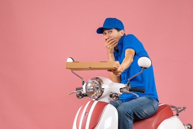 Vista frontal del mensajero preocupado con sombrero sentado en scooter manteniendo orden sobre fondo de melocotón pastel