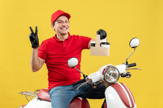 Vista frontal del mensajero seguro hombre vestido con blusa roja y guantes de sombrero en máscara médica entregando orden sentado en scooter sosteniendo órdenes haciendo gesto de victoria