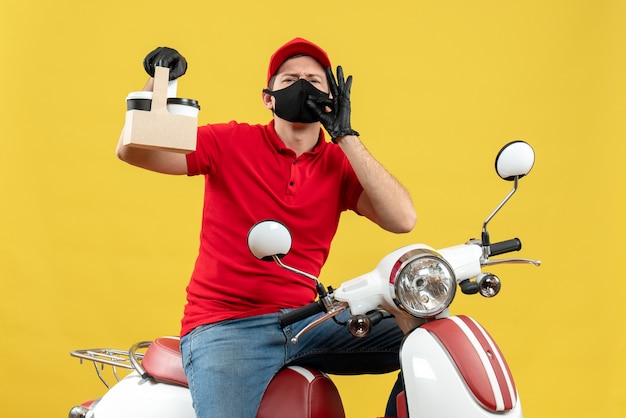 Foto gratuita vista frontal del mensajero seguro hombre vestido con blusa roja y guantes de sombrero en máscara médica sentado en scooter sosteniendo órdenes haciendo gesto ok